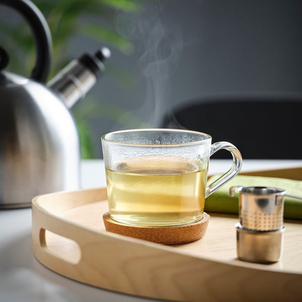 Ein Glasbecher auf einem Holztablett mit dampfendem Tee, neben einem Teekessel und einem Sieb.