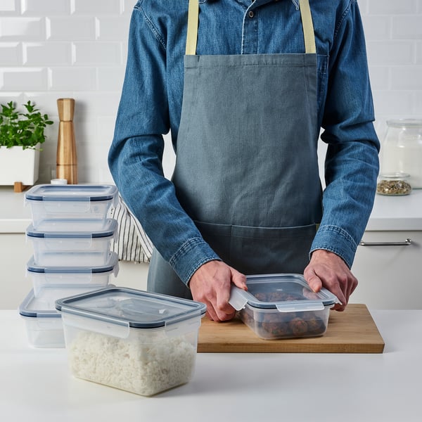 Eine Person in einer Schürze verwendet stapelbare, durchsichtige Lebensmittelbehälter mit Schnappverschluss, um Reste frisch und organisiert zu halten.