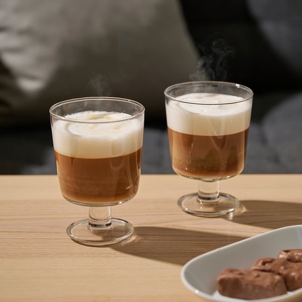 Zwei dampfende Kaffeegetränke in breiten Glastassen auf einem Holztisch. Daneben eine Schüssel mit Schokoladenleckereien.