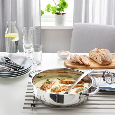 Sauteuse aus Edelstahl auf Untersetzer mit gegrilltem Fisch, Brot und Gläsern auf dem Esstisch am Fenster.