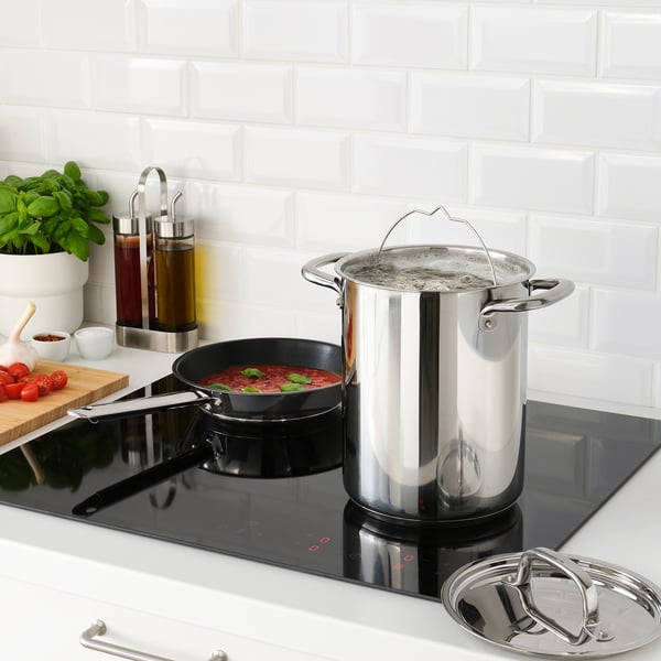 Moderne Küche mit Töpfen und Pfannen aus Edelstahl auf schwarzem Kochfeld, Kochen von Gemüse und Kräutern. IKEA 365+ Kochgeschirr.