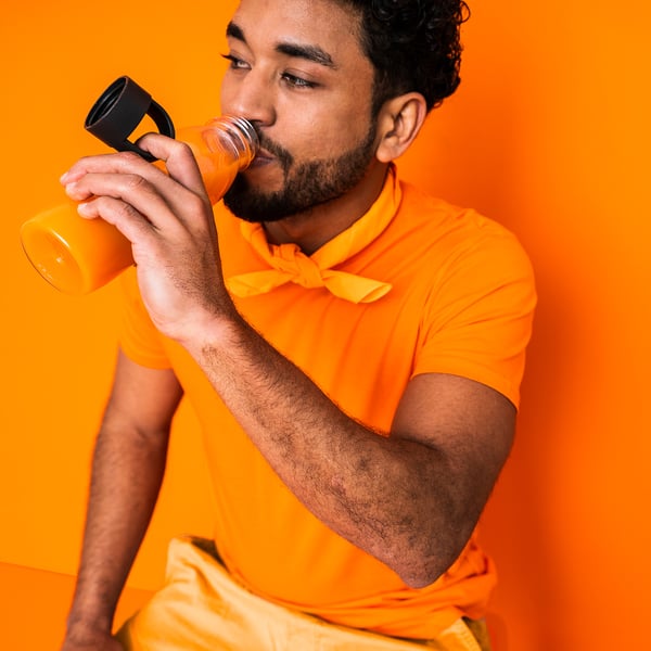 Mann in orangefarbenem Hemd, der aus einer durchsichtigen Flasche mit schwarzem Deckel und Riemen trinkt, vor orangefarbenem Hintergrund.