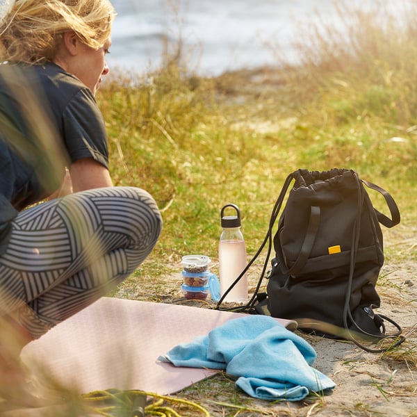 Frau sitzt im Freien mit einer wiederverwendbaren Wasserflasche, einem Rucksack und ein paar Snacks. Die Flasche hat einen Gurt zum einfachen Tragen.