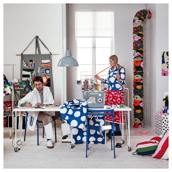Zwei Frauen basteln in einem hellen, farbenfrohen Atelier; der eine näht blauen Polka-Dot-Stoff, der andere skizziert am weißen Tisch; Großes Fenster, lebendige Dekorationen.
