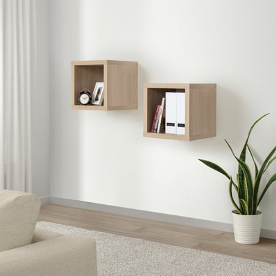KALLAX Wandregale, helles Holz. Kleine, quadratische Würfel nehmen Bücher und Uhr auf. Daneben ein beiger Stuhl und eine Pflanze.