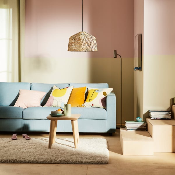 KAPPELAND Rattanlampe hängt über blauem Sofa und wirft warmes Licht, rosa Wände, bunte Kissen und weißen Teppich.