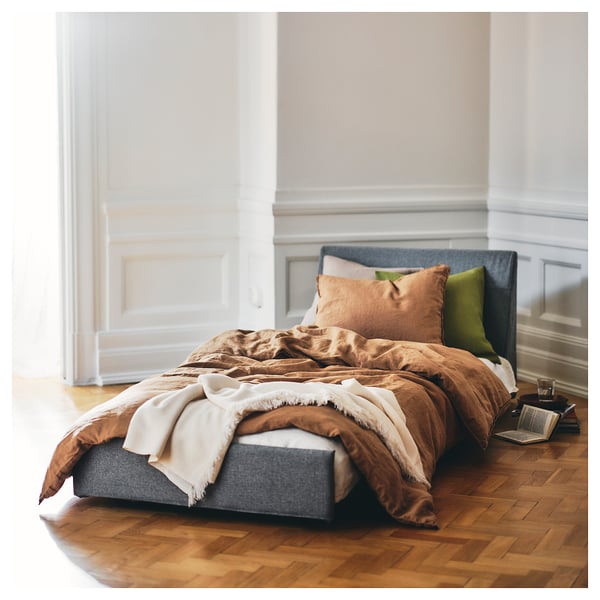Ein gemütliches, kompaktes Bett mit grauem Kopfteil, brauner Steppdecke, weißer Decke, grünem Kissen und Holzboden; KIVIK.