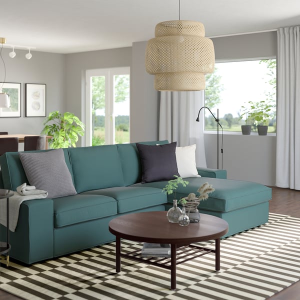 Teal KIVIK Sektionssofa mit gestreiftem Teppich, rundem Couchtisch und Pendelleuchte. Offener Raum mit weißen Vorhängen und Pflanzen.