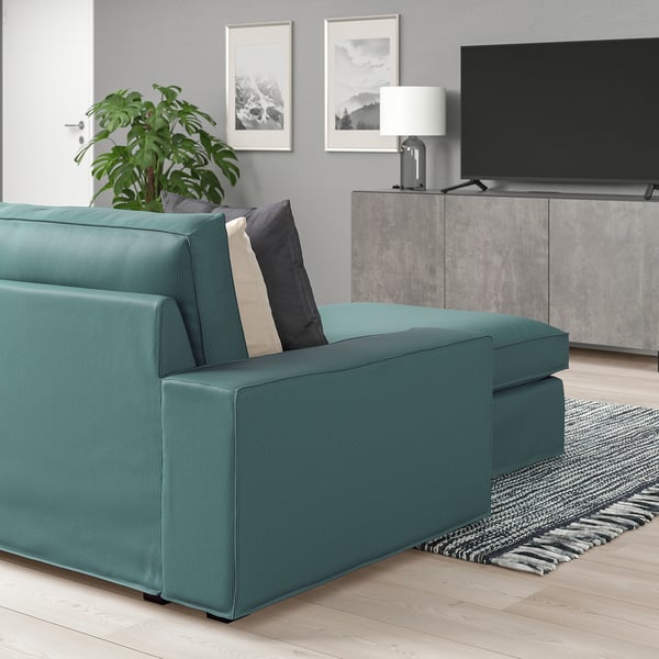 Modernes Wohnzimmer mit blaugrünem KIVIK-Sofa, schwarzen Kissen, grauem Teppich, Pflanze, Fernseher, Lampe und Wandkunst.