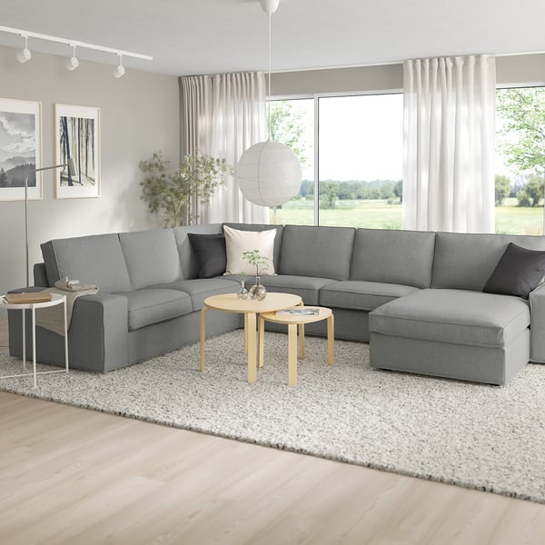Modernes Wohnzimmer mit großem grauem KIVIK-Sofa, zwei Beistelltischen, Teppich und großem Fenster mit Blick ins Grüne.
