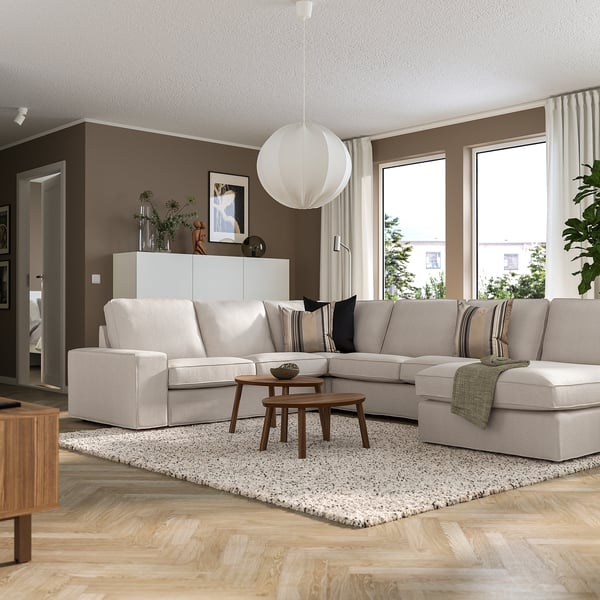 Modernes Wohnzimmer mit großem KIVIK beigem Sektionssofa, das an der braunen Wand abgewinkelt ist, Couchtisch aus Holz und großen Fenstern.