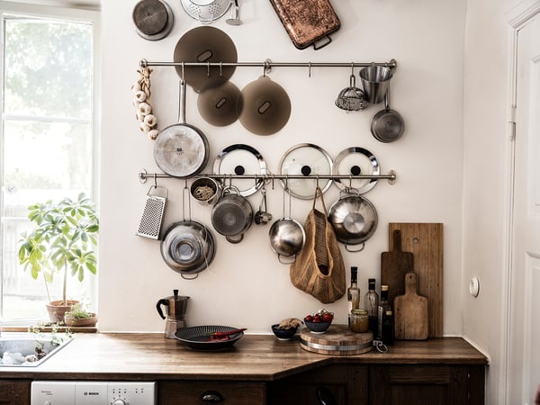 Küche mit Holztheke, hängenden Töpfen, Pfannen, Deckeln, Schneidebrettern und einer Pflanze in der Nähe eines Fensters.