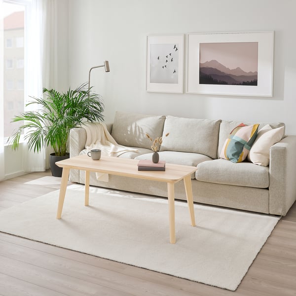 Helles, minimalistisches Wohnzimmer mit weißem KNARDRUP-Teppich, hellgrauem Sofa, Couchtisch aus Holz und Wandkunst. Pflanzen sorgen für Grün.