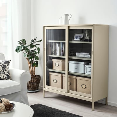 Moderner beiger KVARNVIK-Stauraum im Wohnzimmer mit Glastüren, in dem Bücher und Dekoration ausgestellt sind.