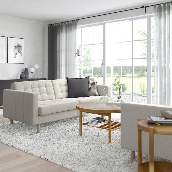 Modernes Wohnzimmer mit grauem LANDSKRONA-Sofa, großem Fenster, Holztischen und Kissen.
