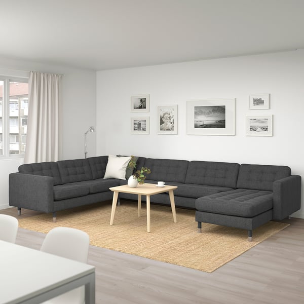 Modernes Wohnzimmer mit großem grauem L-förmigem Sofa, Couchtisch aus Holz und Wandkunst. Heller Holzboden, Juteteppich, weiße Vorhänge.