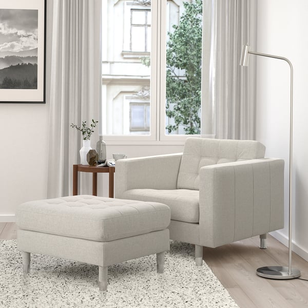 Moderner beiger LANDSKRONA Fußhocker neben dem Sessel, mit Metallbeinen und getuftetem Design. In der Nähe des Fensters mit durchsichtigen Vorhängen und einer Beistelltischvase.