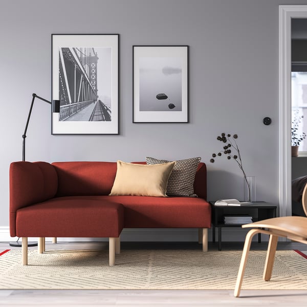 Modernes Wohnzimmer mit rotem modularem LILLEHEM-Sofa, Schwarz-Weiß-Kunst, Beistelltisch, Lampe und Holzstuhl. Graue Wände und gemusterter Teppich.