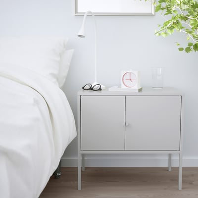 Ein weißer IKEA LIXHULT Aufbewahrungsschrank neben einem Bett. Es ist kompakt, hat zwei Türen und trägt eine Lampe, eine Uhr, ein Glas und eine Vase.