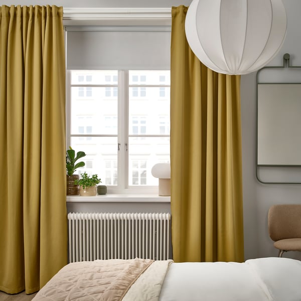 Modernes Schlafzimmer mit MAJGULL Senfvorhängen, weißem Bett, gemütlichem Stuhl und großer Lampe. Vorhänge ragen über Fenster und Bett.