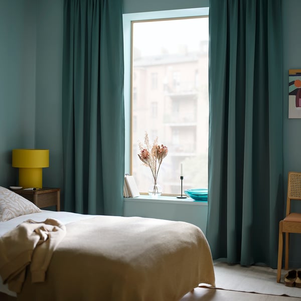Schlafzimmer mit MAJGULL dunkelgrau-türkisfarbenen Raumverdunkelungsvorhängen. Bett zentriert, Lampe links, Fenster hinten.