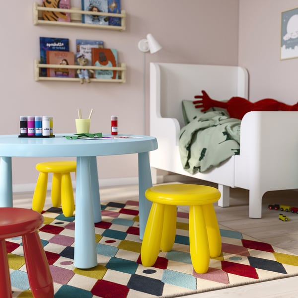 Kinderspielzimmer mit pastellblauem MAMMUT-Tisch, kleinen gelben und roten Hockern, bunter Teppich, weißem Bett mit Bettwäsche, Wandregalen mit Kinderbüchern.