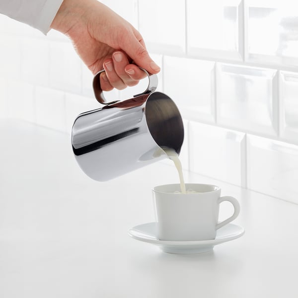 Hand gießt schäumendes Milchkännchen in eine weiße Kaffeetasse auf Untertasse und demonstriert das Aufschäumen von Milch.