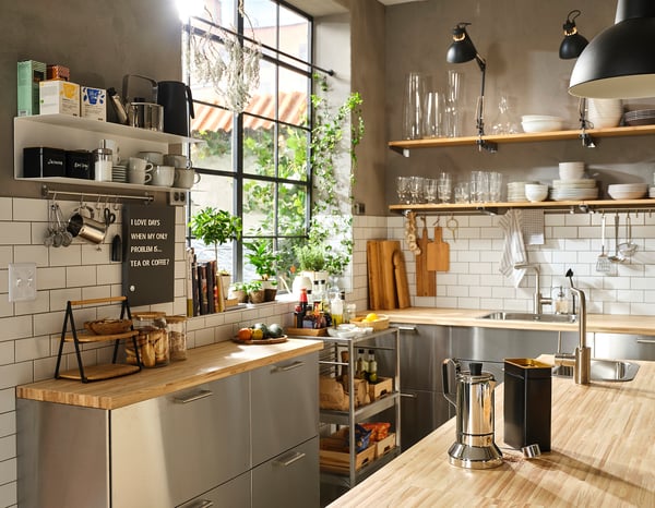 Moderne Küche mit Edelstahlschränken, Holztheken, offenen Regalen und schwarzen Pendelleuchten. Großes Fenster lässt natürliches Licht herein.