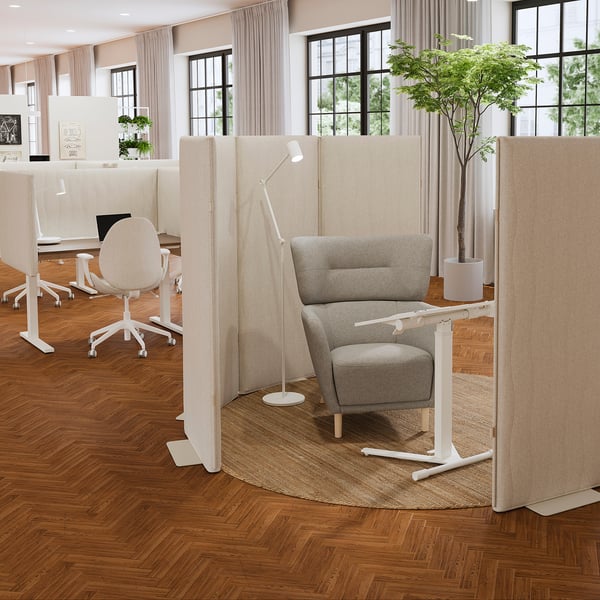 Modernes Büro mit schalldämpfenden Paneelen in MITTZON-Beige, von der Natur inspiriert, zwischen den Schreibtischen. Grauer Sessel, Pflanze, große Fenster mit Vorhängen.