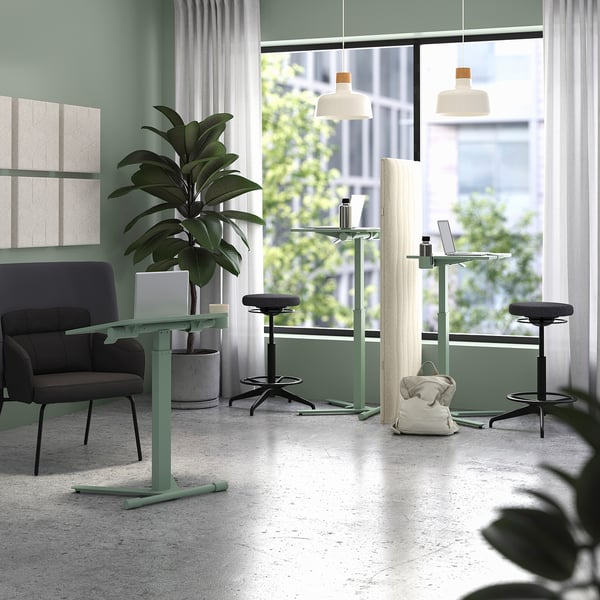 Moderne Büroszene mit grünen MITTZON Laptop-Tischen, schwarzen Hockern und einem grauen Sofa mit großen Fenstern und Pflanzen.