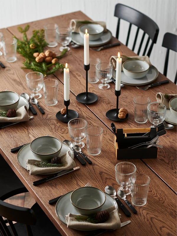 Esstisch aus Holz mit grauen Tellern, Gläsern und Besteck. Kerzen und Grün schmücken das Zentrum. Vier schwarze Stühle drumherum.