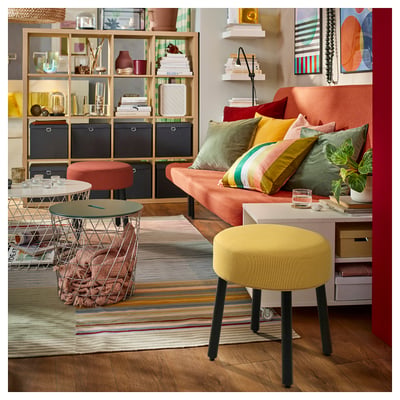 Lebendiges Wohnzimmer mit orangefarbenem Sofa, bunten Kissen, gelbem Hocker, Drahttisch, gestreiftem Teppich und dekorierten Regalen.