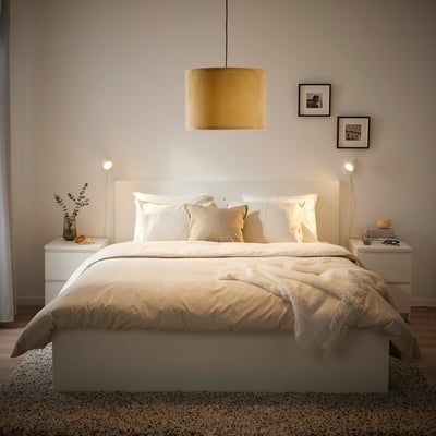 Ein modernes Schlafzimmer mit einem großen weißen Bett, einem gelben Samtlampenschirm und weißen Nachttischen mit Vasen und Nachtlichtern, gerahmte Drucke an der Wand.