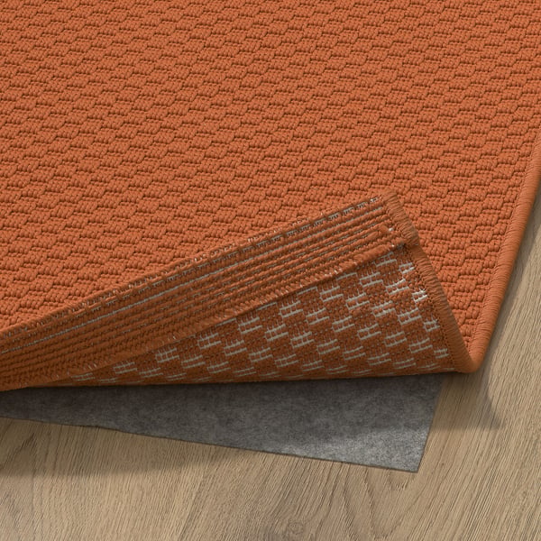 Leuchtend orangefarbener Webteppich auf hellem Hartholzboden mit sichtbarer Rückseite und rutschfester Matte, der sein praktisches Design und seine Unterlage zur Geltung bringt.