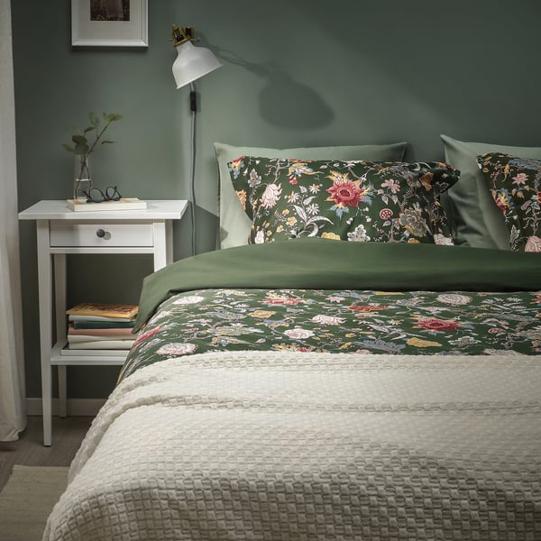 Gemütliches Schlafzimmer mit dunkelgrüner Blumenbettdecke, weißem Nachttisch und Grün.