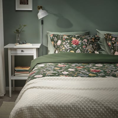 Gemütliches Schlafzimmer mit dunkelgrüner Blumenbettdecke, weißem Nachttisch und Grün.