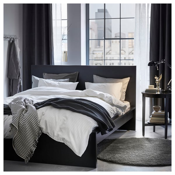 Modernes Schlafzimmer mit großem Fenster, schwarzem Bett mit weißer Bettwäsche, Beistelltisch mit Lampe. Elegantes, minimalistisches Design.