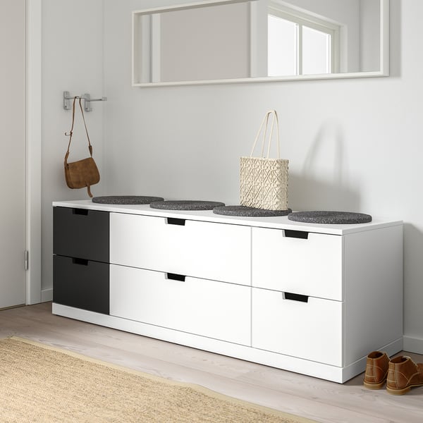 Moderne weiße NORDLI-Kommode mit sechs Schubladen mit schwarzem Griff, in der sich eine geflochtene Tasche unter einem Spiegel befindet.