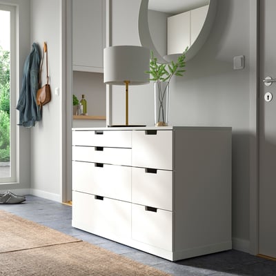Moderner Flur mit weißer NORDLI Truhe, Lampe, Spiegel und Kleiderhaken. Die Kommode hat vier Schubladen, die an einer Wand in der Nähe einer Tür platziert sind.