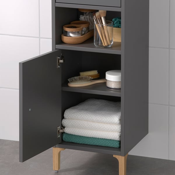Hoher grauer Badezimmerschrank mit offenem und geschlossenem Stauraum. Bietet Platz für Handtücher, Toilettenartikel und Pflegewerkzeuge. Muss aus Sicherheitsgründen an der Wand montiert werden.