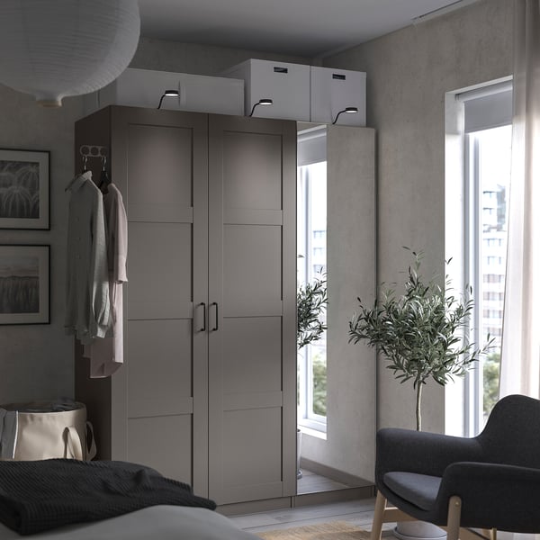 Moderner grauer PAX-Kleiderschrank im Schlafzimmer mit Spiegeltüren, offenem Kleiderschrank, hängender Kleidung, grauem Sessel, Beistelltisch, Fenster und Zimmerpflanze.