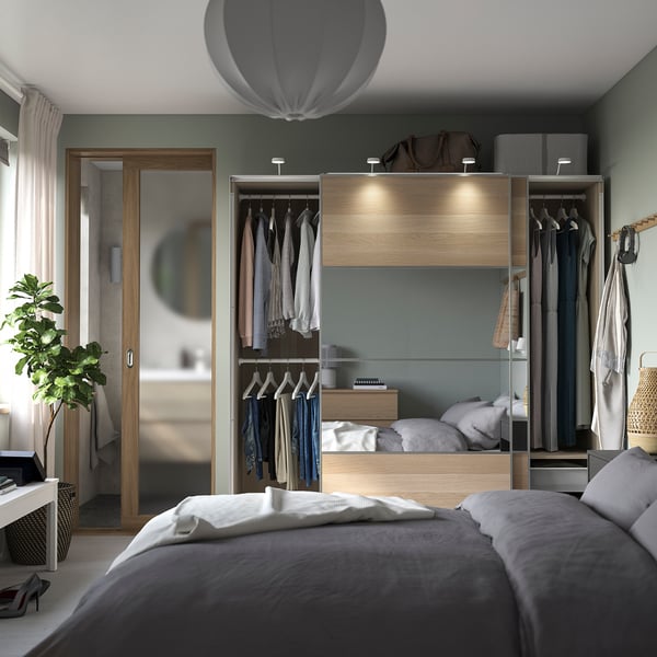 Modernes Schlafzimmer mit PAX-Kleiderschrank, Schiebetüren, organisierter Kleidung, integrierter Beleuchtung, grauen Laken, weißen Kissen, Holzakzenten und grünen Wänden.