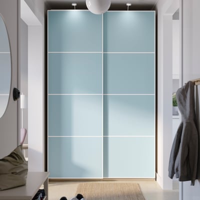 Ein hoher, hellblauer PAX-Kleiderschrank mit Schiebetüren. Der Kleiderschrank ist größer als der nahe gelegene Spiegel und die Garderobe und verfügt über ein schlankes, modernes Design.