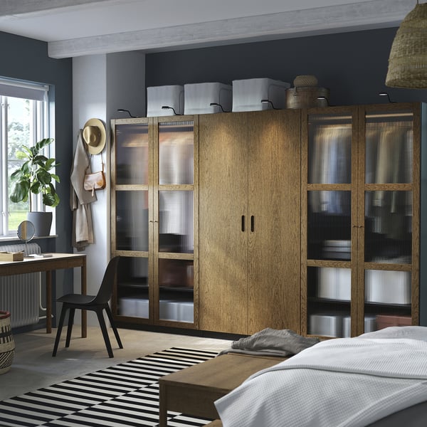 Modernes Schlafzimmer mit PAX-Kleiderschrank: helles Holz, Glastüren, verschiedene Regale und Körbe, schwarzer Stuhl, gestreifter Teppich, Topfpflanze.