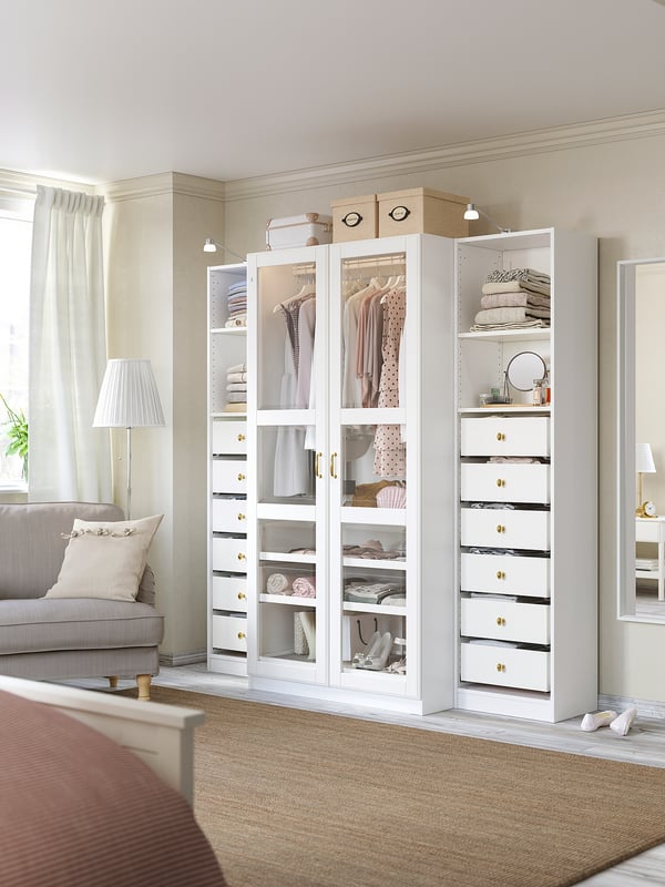 Modernes Schlafzimmer mit weißem PAX-Kleiderschrank mit Glas und massiven Schubladen, in dem sich Kleidung und Accessoires neben einem grauen Sofa und einem Teppich befinden.