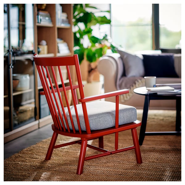 Roter Spindelsessel mit Kissen im gemütlichen Wohnzimmer. Holzrahmen, kleiner als die Couch, mit leichtem Kissen. Enthält ein Bücherregal, Pflanzen und einen warmen Teppich.