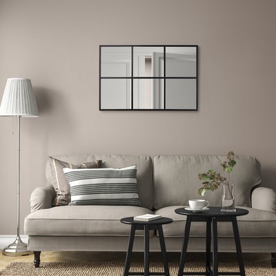 Minimalistisches Wohnzimmer mit RAGGARÖN-Spiegel über grauem Sofa, gestreiften Kissen, kleinen Tischen mit Buch, Tasse und Vase sowie einer hohen Stehlampe.