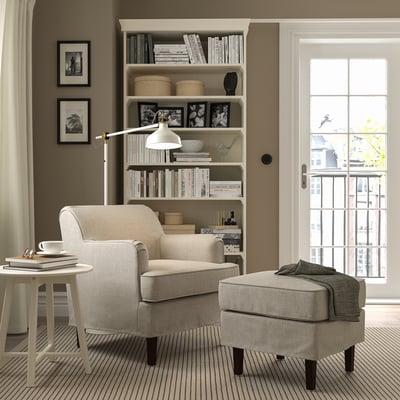 Beige Sessel & Ottomane, Bücherregal, Beistelltisch mit Lampe, großes Fenster.