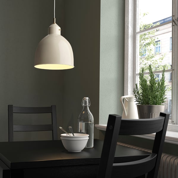 Eine kleine weiße RÖDFLIK-Lampe hängt in der Nähe eines Fensters und beleuchtet einen dunklen Tisch mit Stühlen und Dekoration.