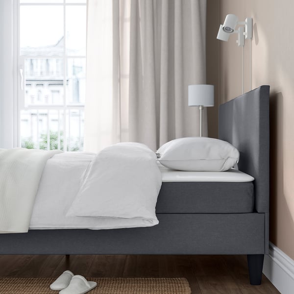 Bett mit grau gepolstertem Kopfteil, weißer Bettwäsche, Wandleuchte und Hausschuhen auf einer Matte, an einem großen Fenster gelegen.
